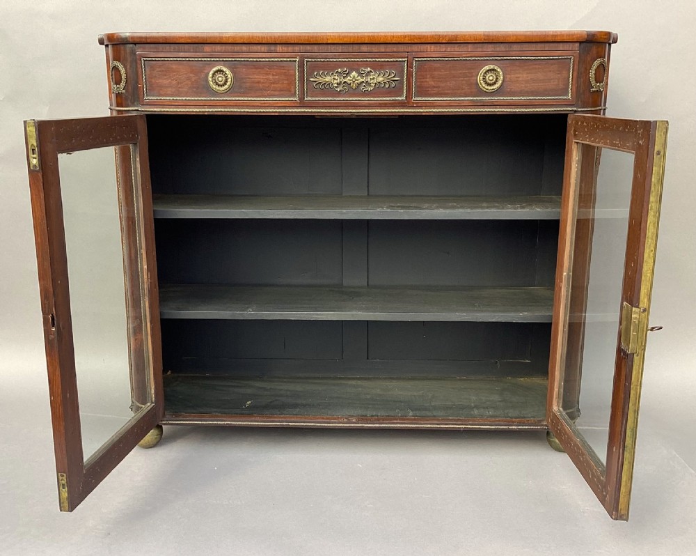 A Regency period dark rosewood side cabinet featuring gilt brass mouldings, decorative wreath appliques, two drawers, glass doors, and brass ball feet. View XII