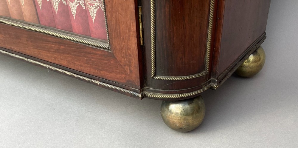 A Regency period dark rosewood side cabinet featuring gilt brass mouldings, decorative wreath appliques, two drawers, glass doors, and brass ball feet. View XXII