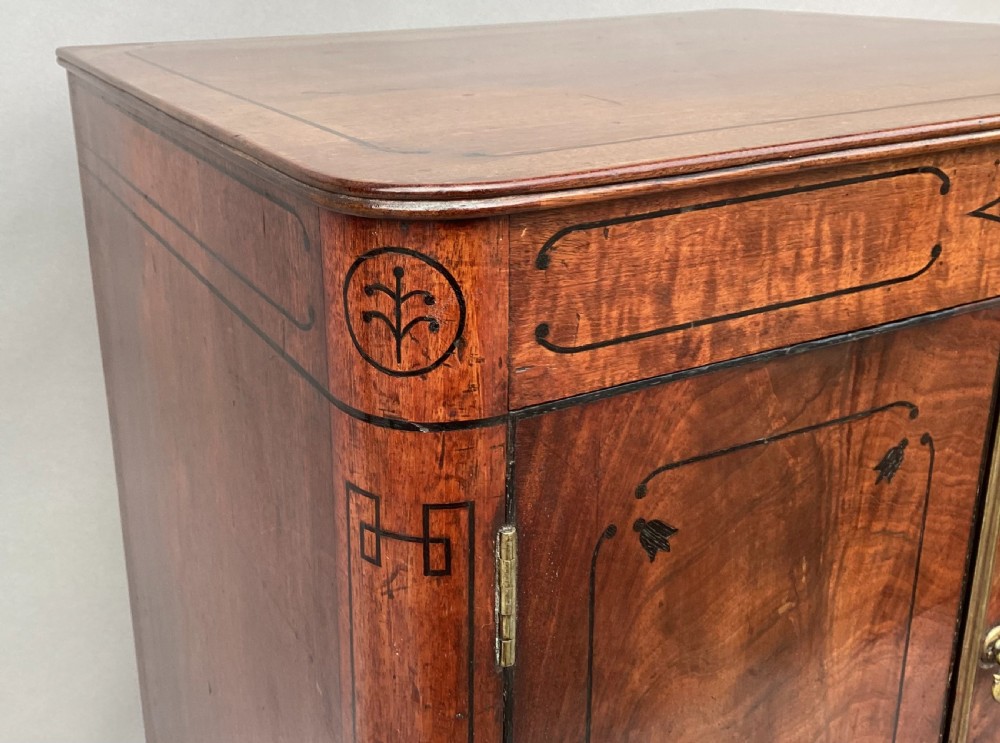 A Regency flame mahogany pedestal cabinet featuring dark ebony inlay stringing, upper double doors, a lower cupboard door, and brass paw feet. View IX