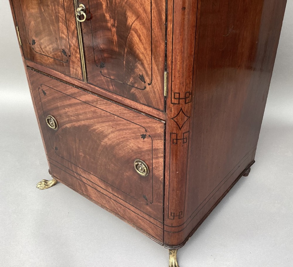 A Regency flame mahogany pedestal cabinet featuring dark ebony inlay stringing, upper double doors, a lower cupboard door, and brass paw feet. View XVIII