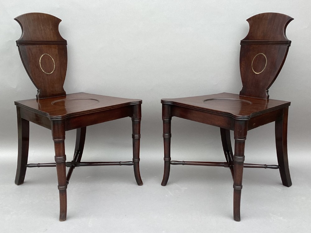 A pair of George III mahogany hall chairs with shield-shaped backs featuring circular brass inlays, dished wooden seats, and turned legs.