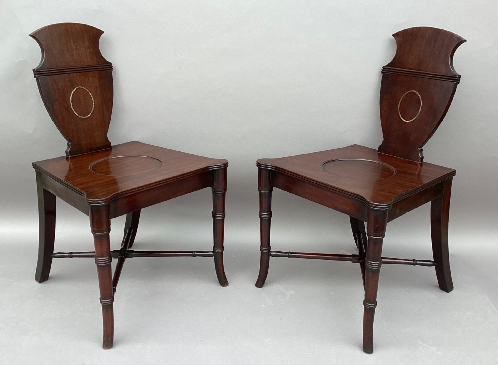A pair of George III mahogany hall chairs with shield-shaped backs featuring circular brass inlays, dished wooden seats, and turned legs. View III