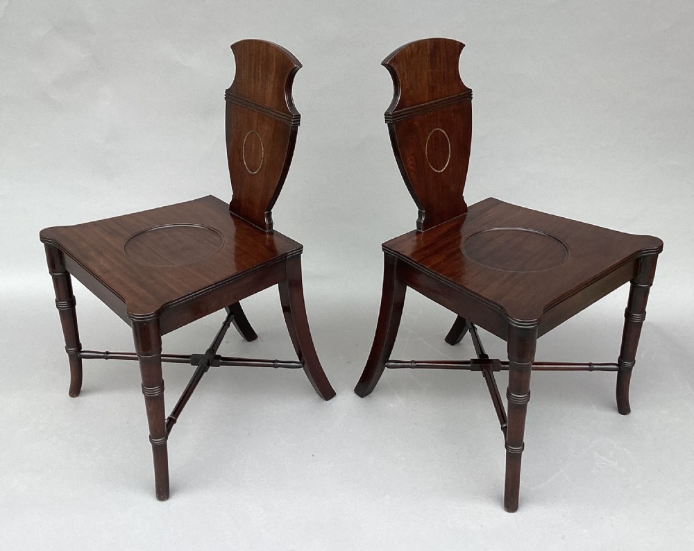 A pair of George III mahogany hall chairs with shield-shaped backs featuring circular brass inlays, dished wooden seats, and turned legs. View VIII