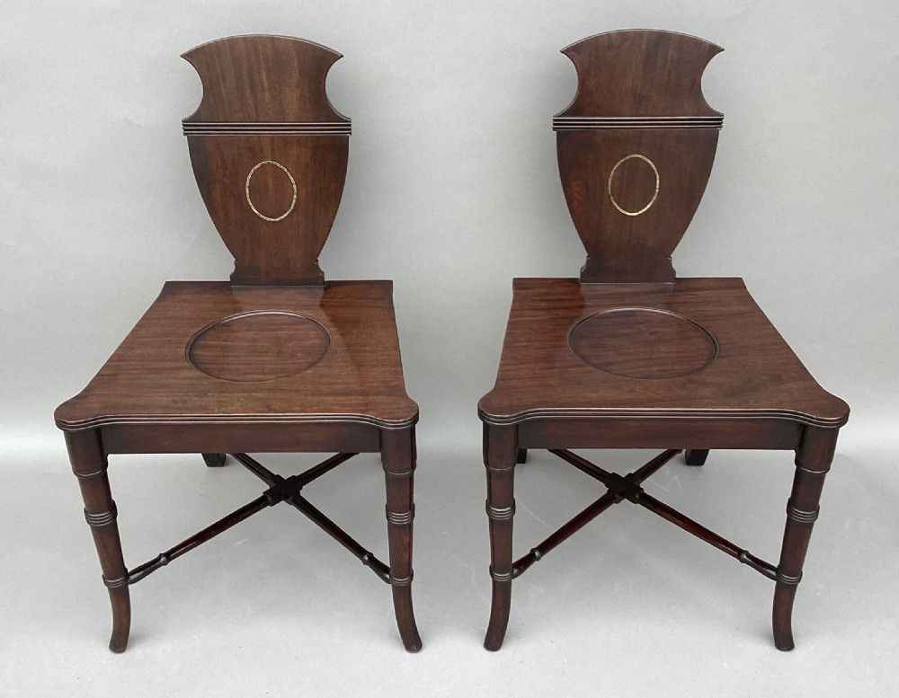 A pair of George III mahogany hall chairs with shield-shaped backs featuring circular brass inlays, dished wooden seats, and turned legs. View X
