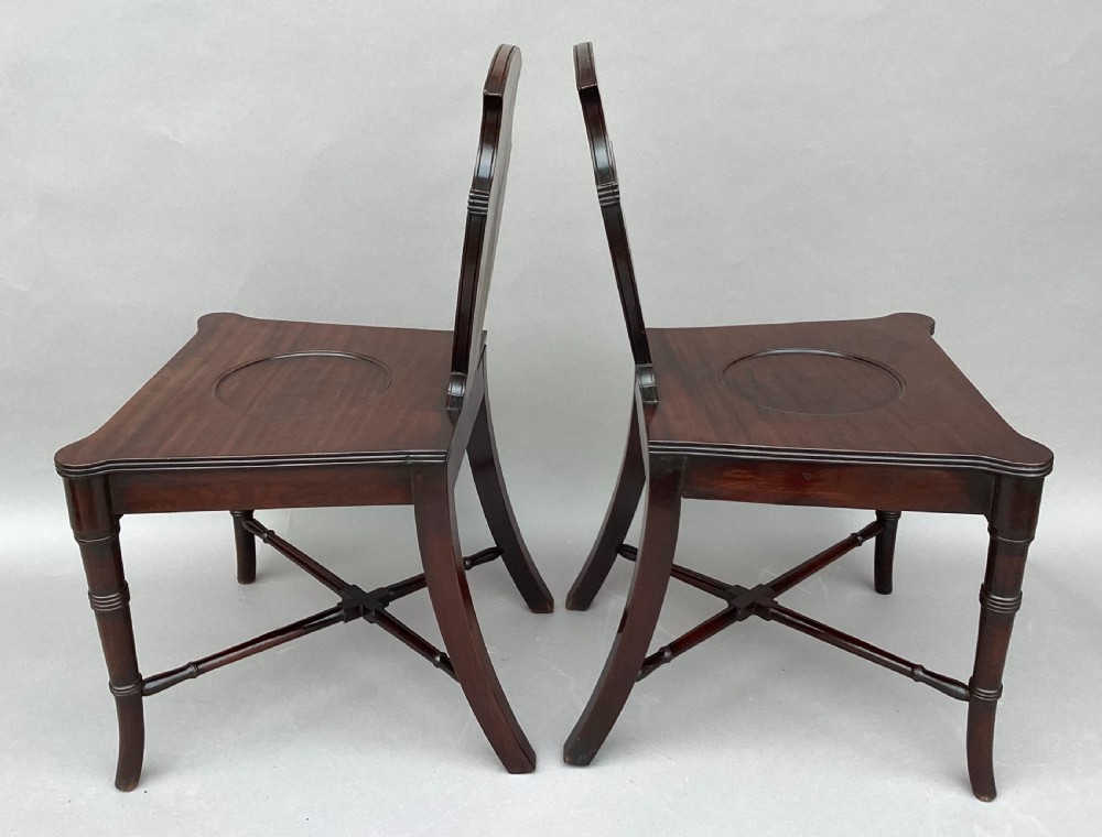 A pair of George III mahogany hall chairs with shield-shaped backs featuring circular brass inlays, dished wooden seats, and turned legs. View XII