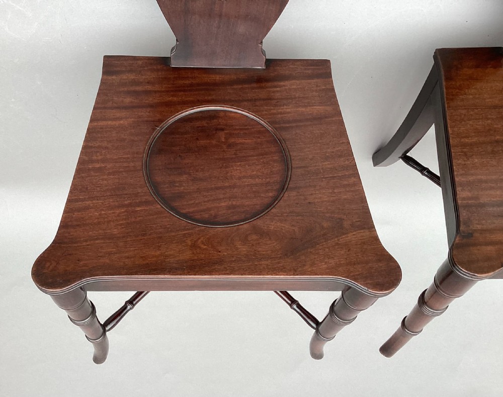 A pair of George III mahogany hall chairs with shield-shaped backs featuring circular brass inlays, dished wooden seats, and turned legs. View XVI