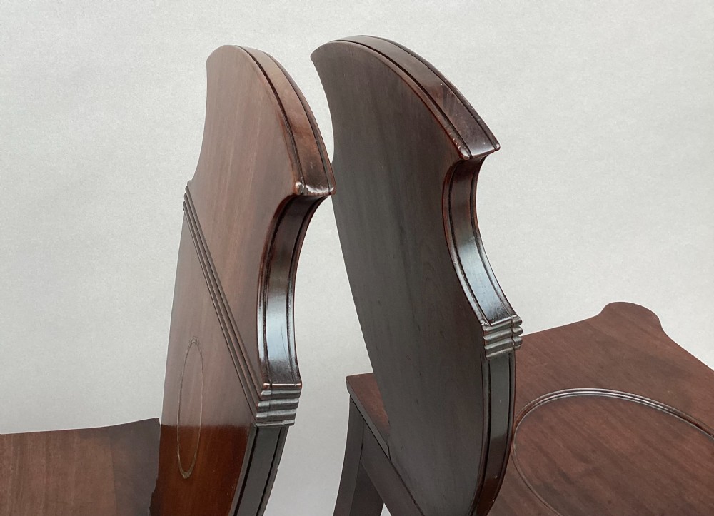 A pair of George III mahogany hall chairs with shield-shaped backs featuring circular brass inlays, dished wooden seats, and turned legs. View XVIII