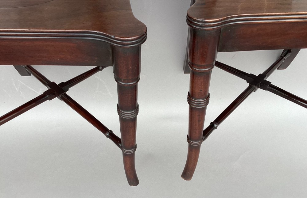 A pair of George III mahogany hall chairs with shield-shaped backs featuring circular brass inlays, dished wooden seats, and turned legs. View XIX
