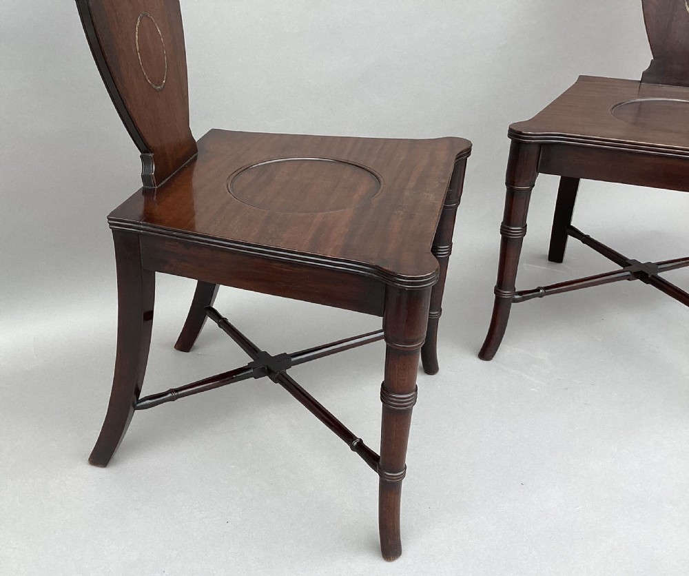 A pair of George III mahogany hall chairs with shield-shaped backs featuring circular brass inlays, dished wooden seats, and turned legs. View XXI