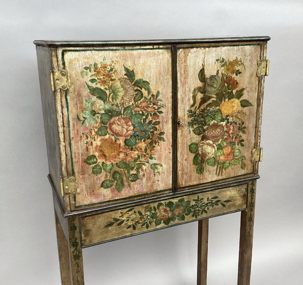 An eighteenth-century hand-painted wooden cabinet on a tall stand, featuring floral artwork across the twin doors, side panels, and apron, mounted with brass hinges. View XVIII