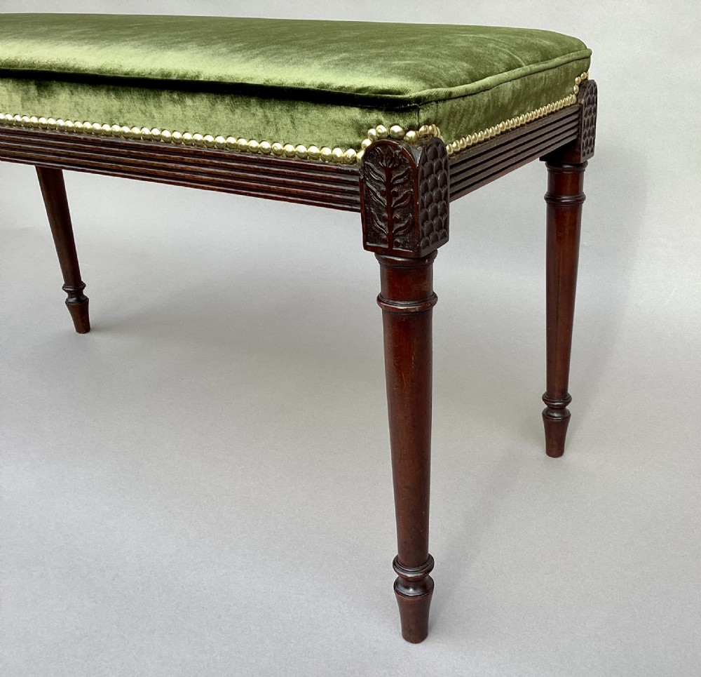 A rectangular George III mahogany window seat featuring sage green velvet upholstery with brass studding, reeded rails, carved corner blocks, and tapered turned legs. View VI