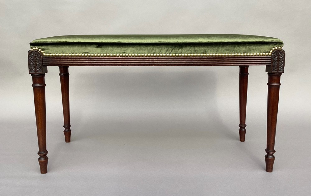 A rectangular George III mahogany window seat featuring sage green velvet upholstery with brass studding, reeded rails, carved corner blocks, and tapered turned legs. View VIII