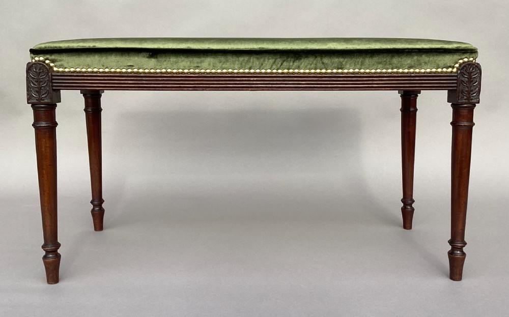 A rectangular George III mahogany window seat featuring sage green velvet upholstery with brass studding, reeded rails, carved corner blocks, and tapered turned legs. View IX
