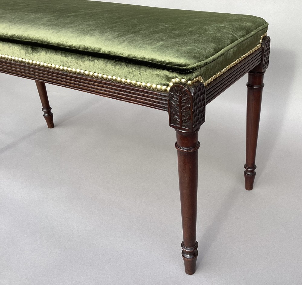 A rectangular George III mahogany window seat featuring sage green velvet upholstery with brass studding, reeded rails, carved corner blocks, and tapered turned legs. View X