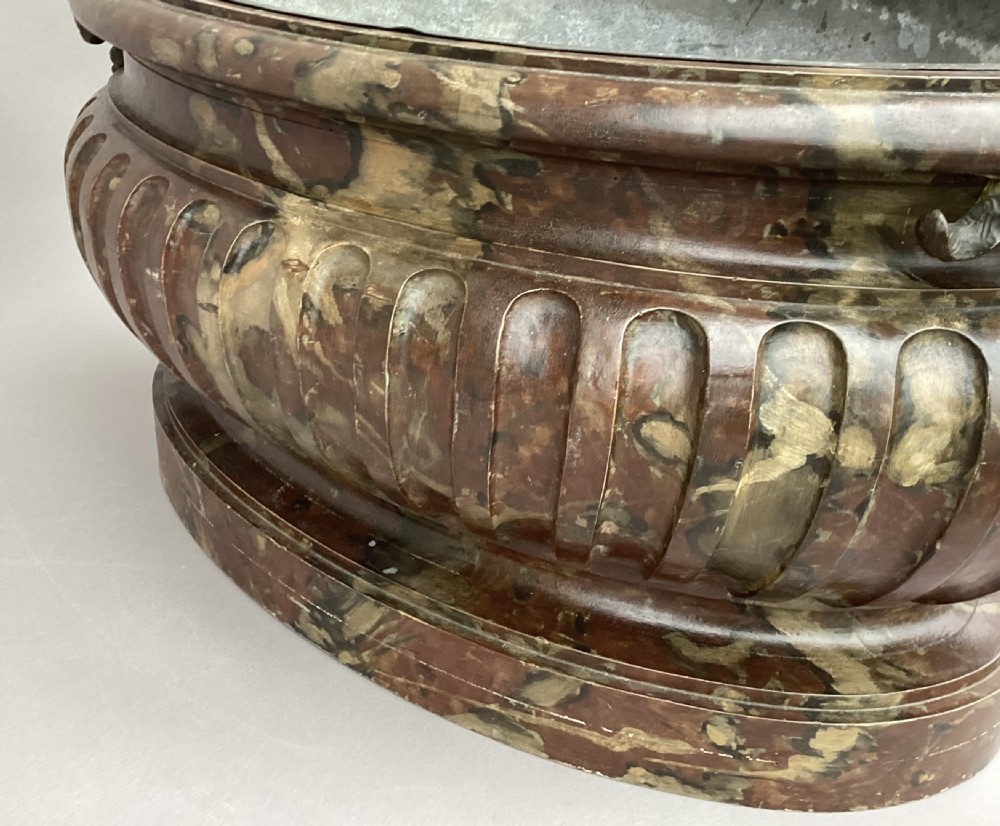 A Regency style oval wine cistern featuring a faux marble painted body with fluted decoration, cast bronze ram head handles, and an internal metal liner. View V