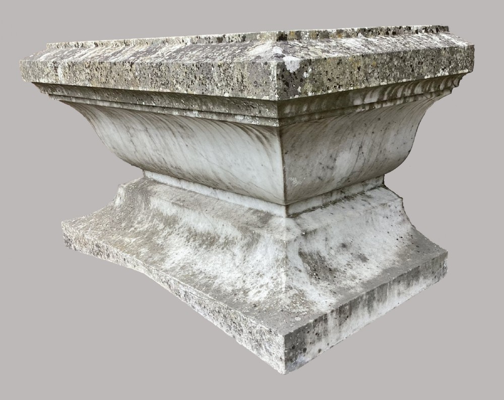 A late 18th-century Neoclassical Carrara marble cistern featuring a curved basin, flared top rim moulding, and a stepped rectangular base with a weathered stone patina. View II