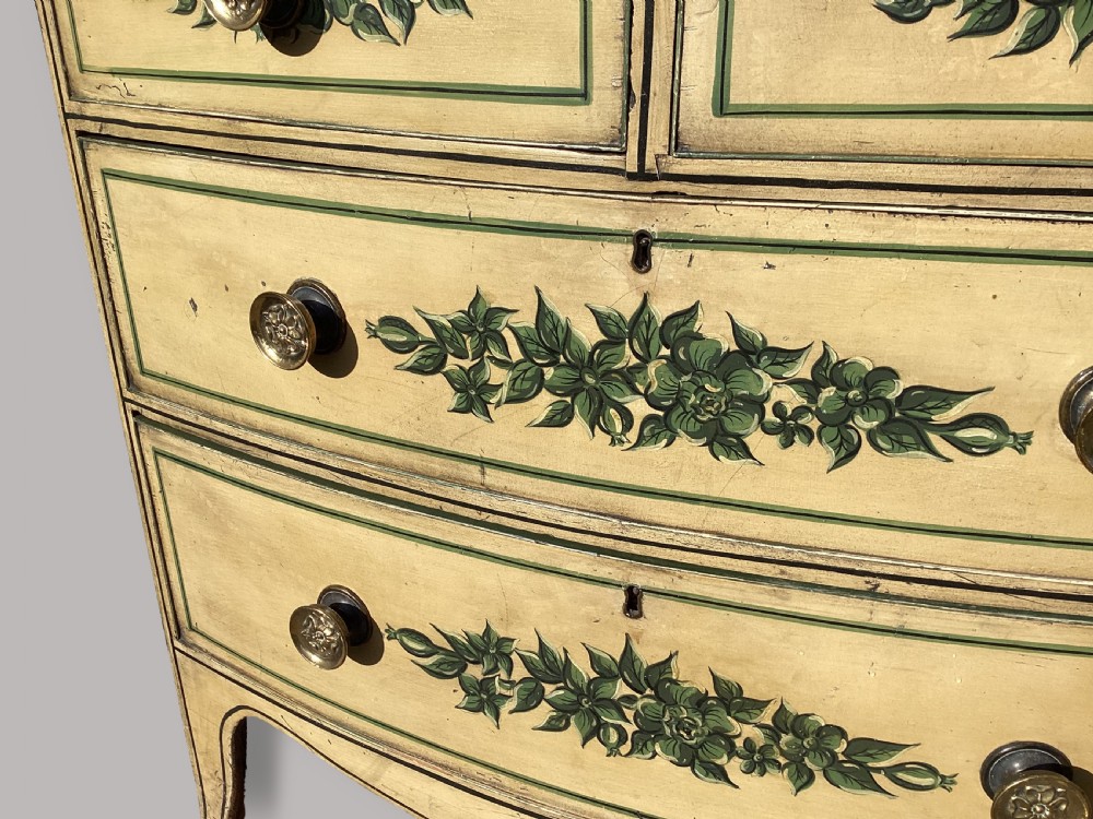 An antique Regency bow-fronted chest of drawers finished in cream paint with hand-painted green floral swags and brass knobs. View V