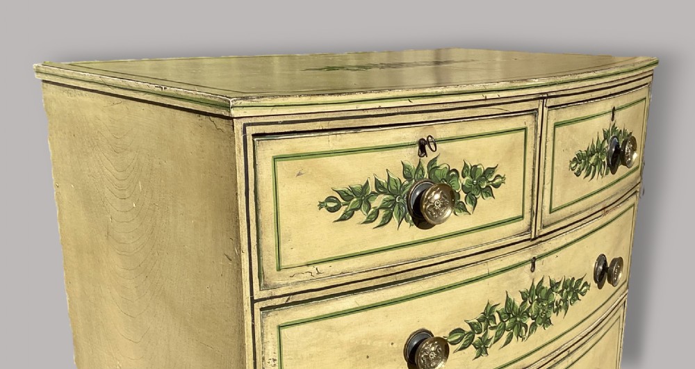 An antique Regency bow-fronted chest of drawers finished in cream paint with hand-painted green floral swags and brass knobs. View VII