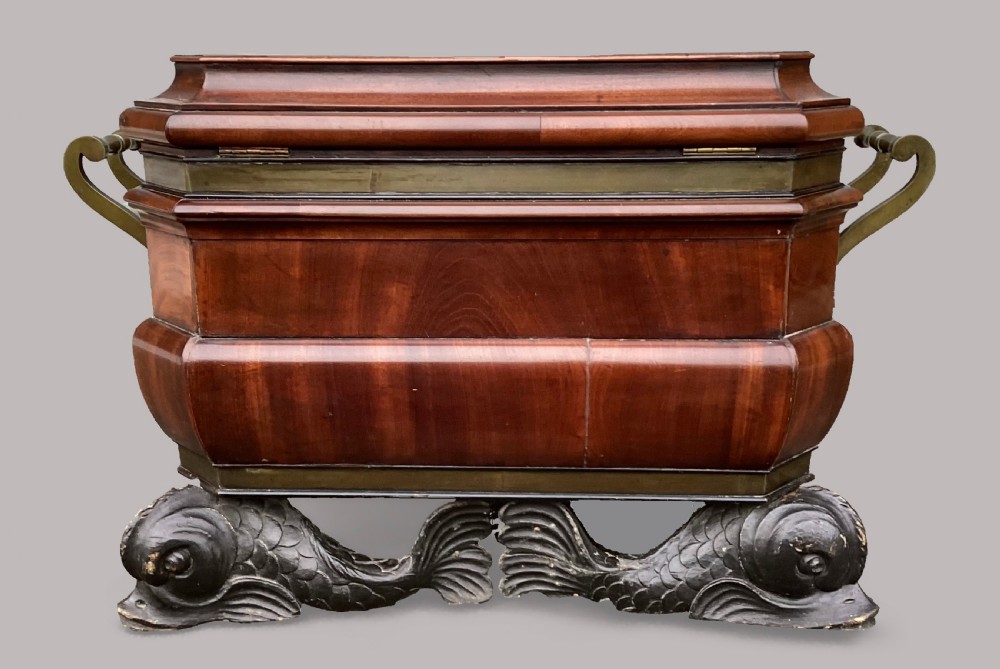A Regency period mahogany wine cellarette of sarcophagus form featuring brass banding, carrying handles, and carved dolphin feet. View V