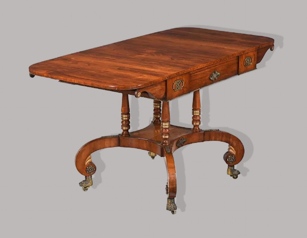 A Regency period rosewood drop-leaf sofa table featuring frieze drawers, turned columns, gilt brass mounts, and splayed legs with brass castors.