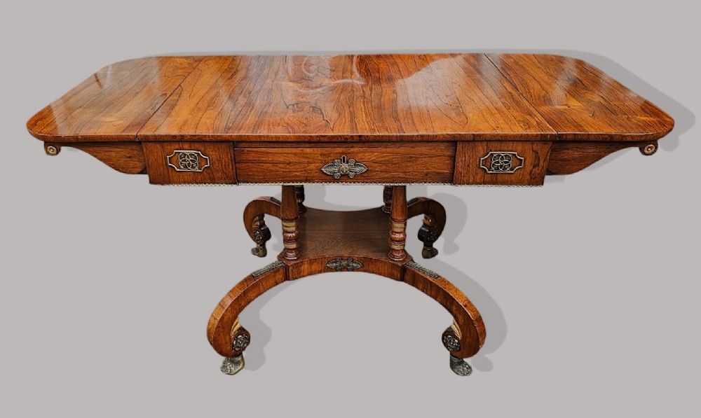 A Regency period rosewood drop-leaf sofa table featuring frieze drawers, turned columns, gilt brass mounts, and splayed legs with brass castors. View V