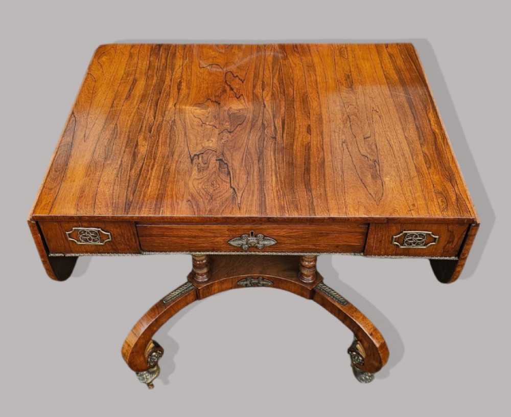 A Regency period rosewood drop-leaf sofa table featuring frieze drawers, turned columns, gilt brass mounts, and splayed legs with brass castors. View VI