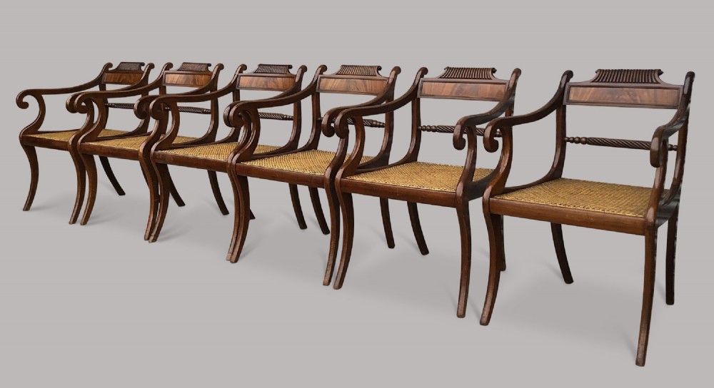 A set of six Regency mahogany open armchairs featuring woven cane seats, curved scroll arms, reeded top rails, and sabre legs.