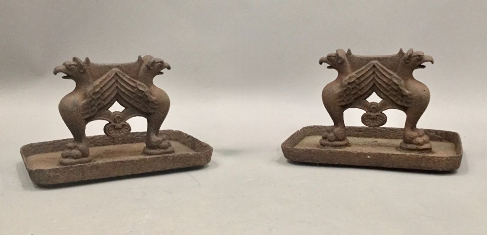 A pair of antique cast iron boot scrapers featuring sculpted griffin side supports and rectangular drip trays with an oxidised surface.