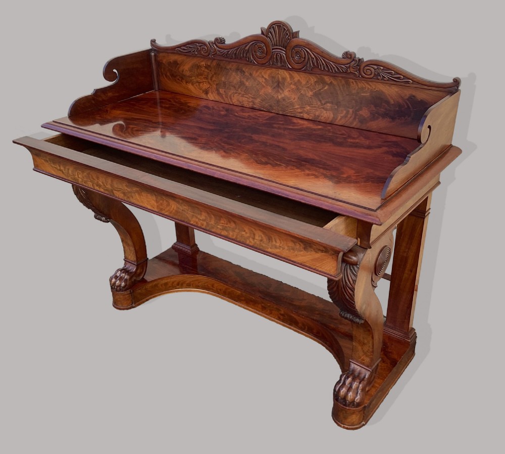 A Regency mahogany console table featuring a carved scroll cresting backboard, flame mahogany veneers, carved acanthus leaf legs, and paw feet on a plinth base. View III
