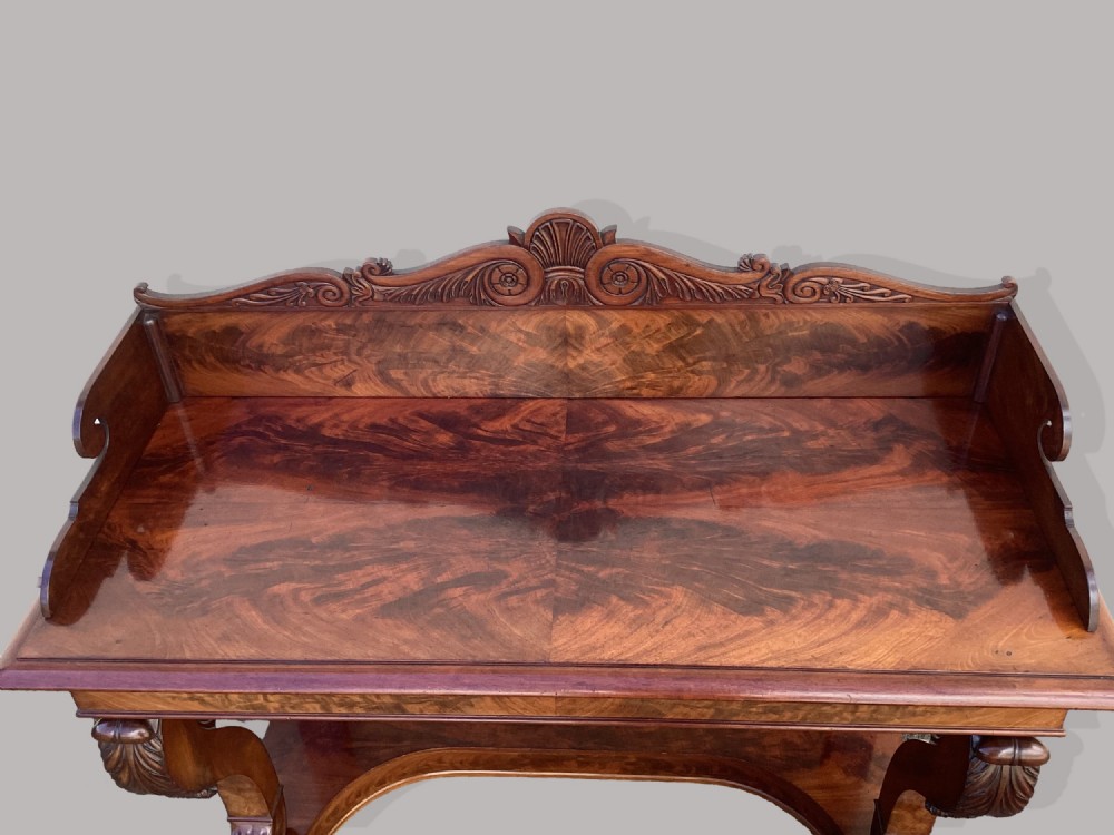 A Regency mahogany console table featuring a carved scroll cresting backboard, flame mahogany veneers, carved acanthus leaf legs, and paw feet on a plinth base. View IV