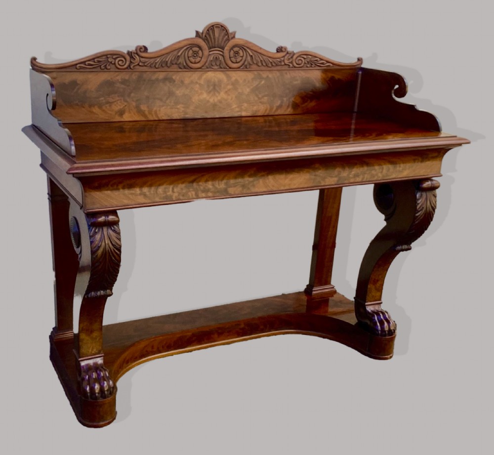 A Regency mahogany console table featuring a carved scroll cresting backboard, flame mahogany veneers, carved acanthus leaf legs, and paw feet on a plinth base. View VI