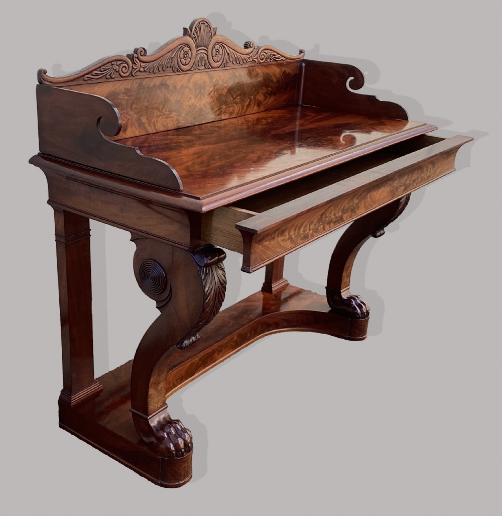 A Regency mahogany console table featuring a carved scroll cresting backboard, flame mahogany veneers, carved acanthus leaf legs, and paw feet on a plinth base. View VII