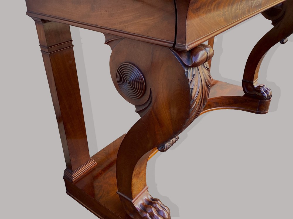 A Regency mahogany console table featuring a carved scroll cresting backboard, flame mahogany veneers, carved acanthus leaf legs, and paw feet on a plinth base. View VIII