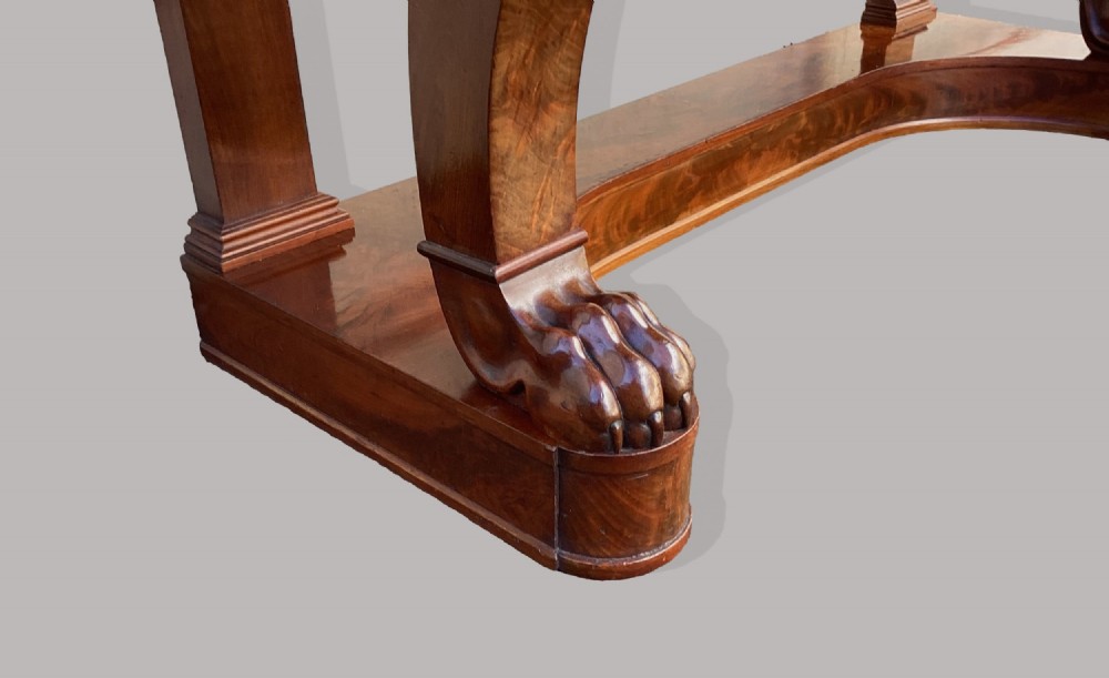 A Regency mahogany console table featuring a carved scroll cresting backboard, flame mahogany veneers, carved acanthus leaf legs, and paw feet on a plinth base. View IX