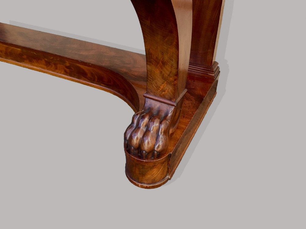 A Regency mahogany console table featuring a carved scroll cresting backboard, flame mahogany veneers, carved acanthus leaf legs, and paw feet on a plinth base. View X