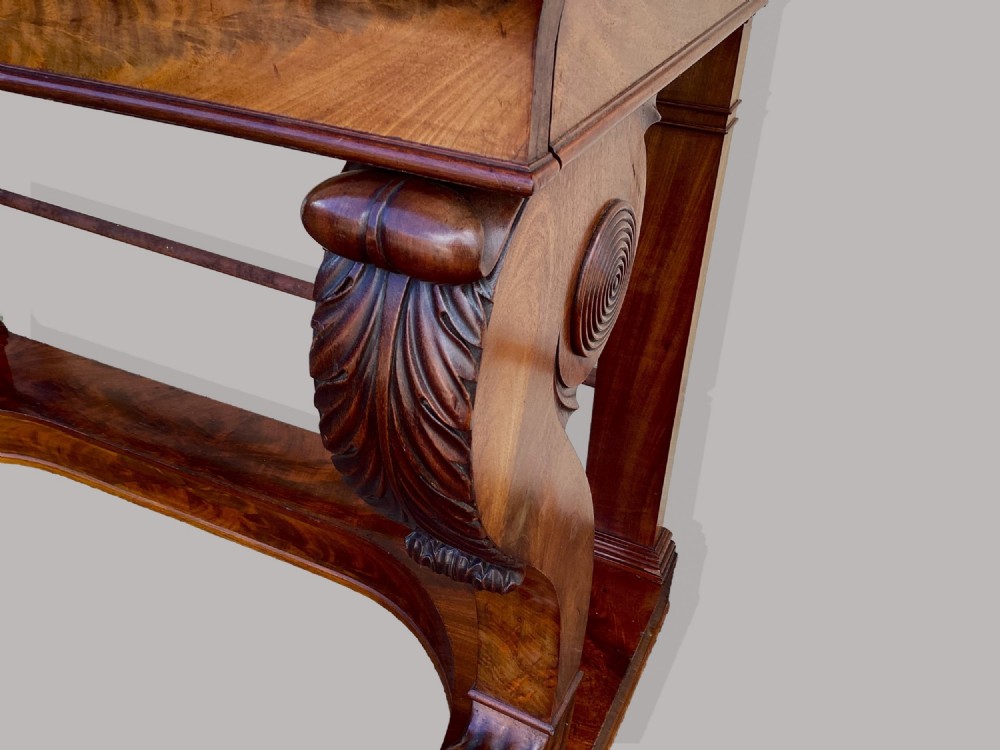 A Regency mahogany console table featuring a carved scroll cresting backboard, flame mahogany veneers, carved acanthus leaf legs, and paw feet on a plinth base. View XI