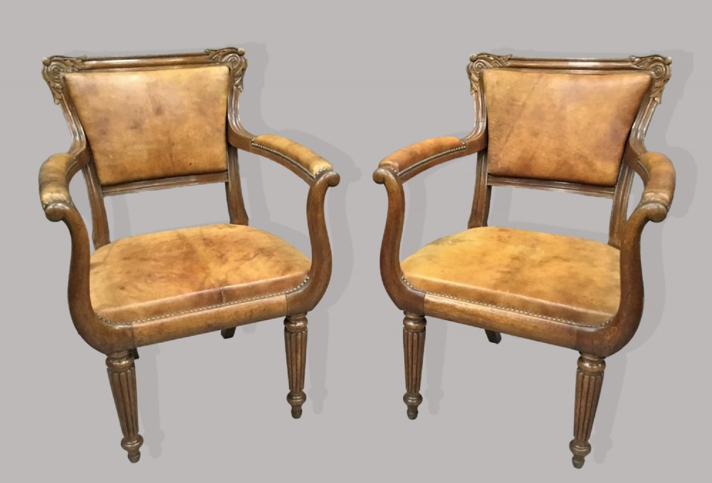 A pair of Regency oak armchairs featuring tan leather upholstery, carved scrolled cresting rails, shaped armrests, and reeded front legs with brass stud trim.