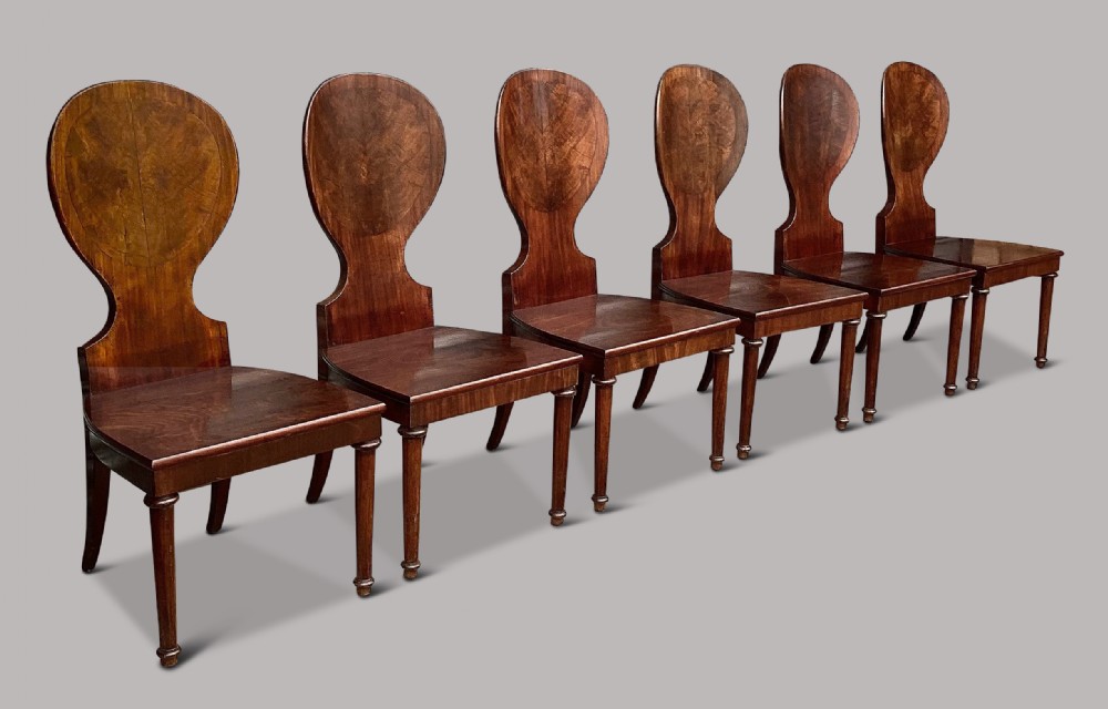 A set of six George III mahogany hall chairs featuring rounded cartouche-shaped backrests, solid wooden seats, and turned front legs.