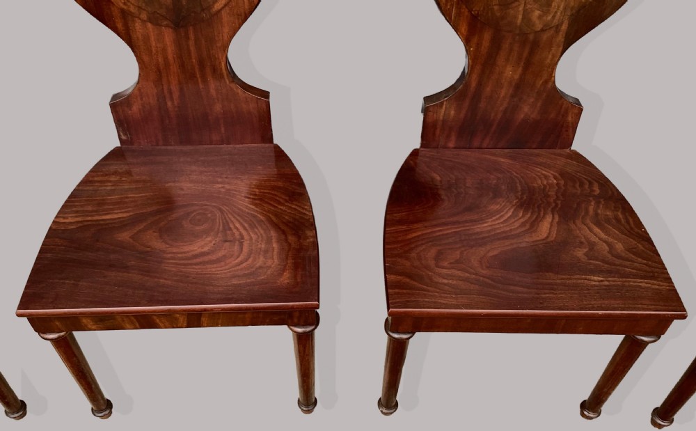A set of six George III mahogany hall chairs featuring rounded cartouche-shaped backrests, solid wooden seats, and turned front legs. View IV