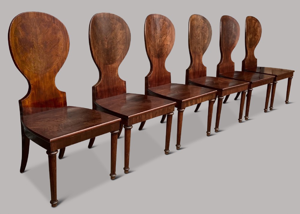 A set of six George III mahogany hall chairs featuring rounded cartouche-shaped backrests, solid wooden seats, and turned front legs. View V