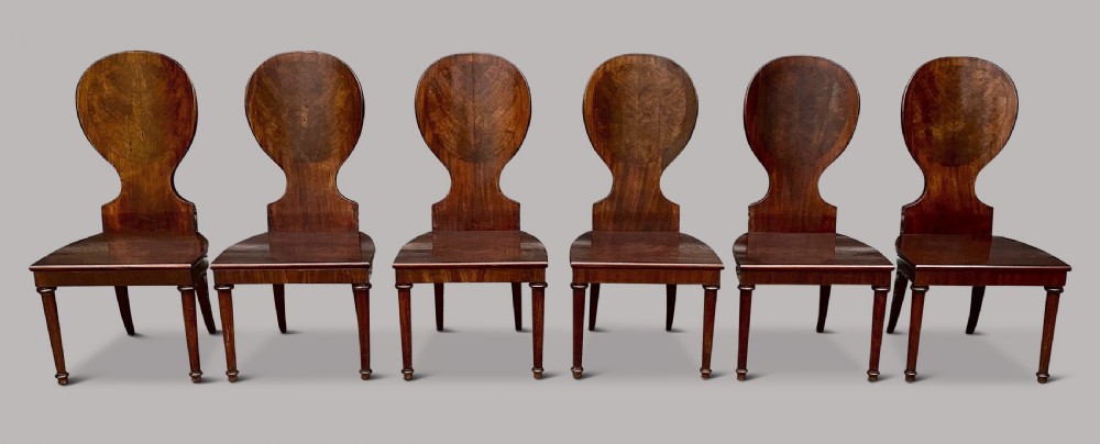 A set of six George III mahogany hall chairs featuring rounded cartouche-shaped backrests, solid wooden seats, and turned front legs. View VIII