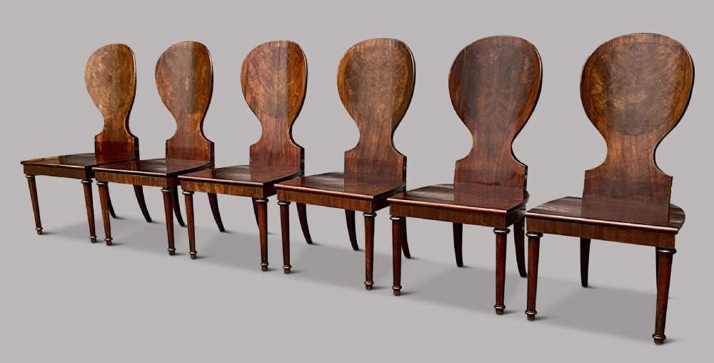 A set of six George III mahogany hall chairs featuring rounded cartouche-shaped backrests, solid wooden seats, and turned front legs. View IX