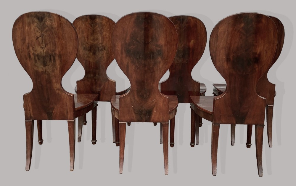 A set of six George III mahogany hall chairs featuring rounded cartouche-shaped backrests, solid wooden seats, and turned front legs. View XX