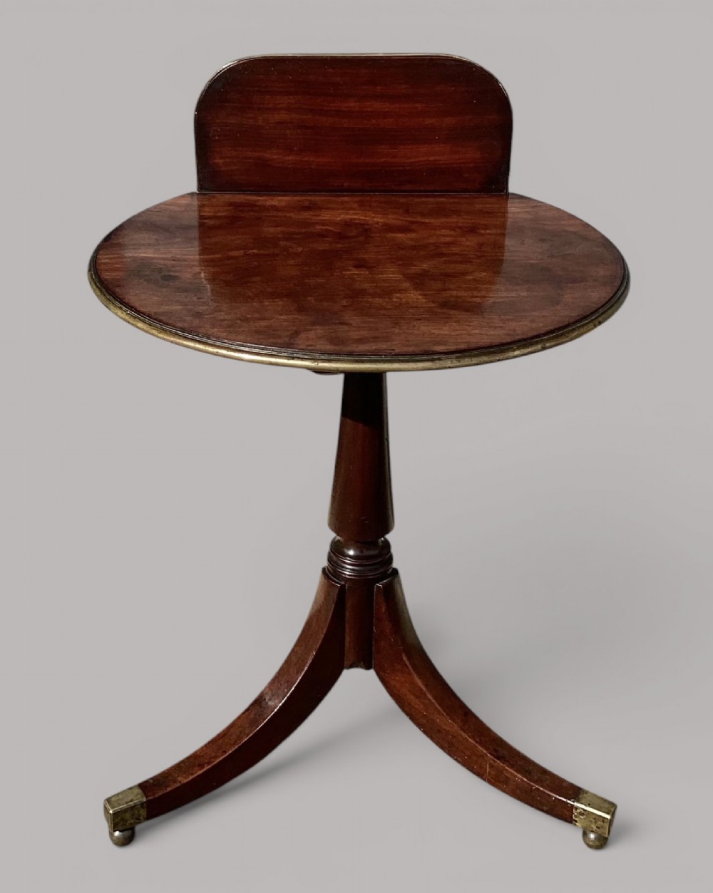 A George III mahogany tripod occasional table featuring a raised back panel, brass moulding on the circular top, a turned central column, and three curved legs ending in brass caps.