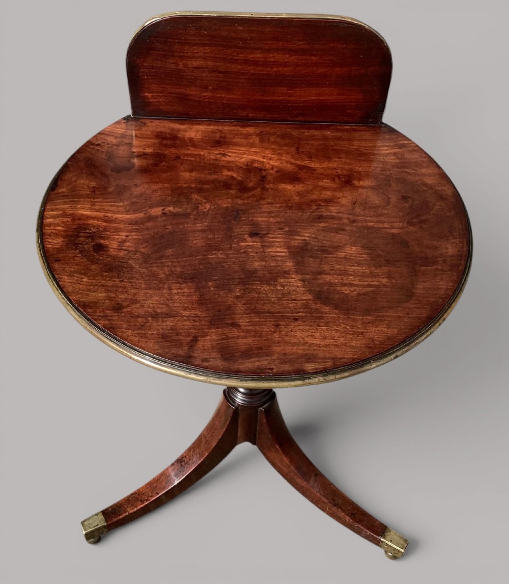 A George III mahogany tripod occasional table featuring a raised back panel, brass moulding on the circular top, a turned central column, and three curved legs ending in brass caps. View II