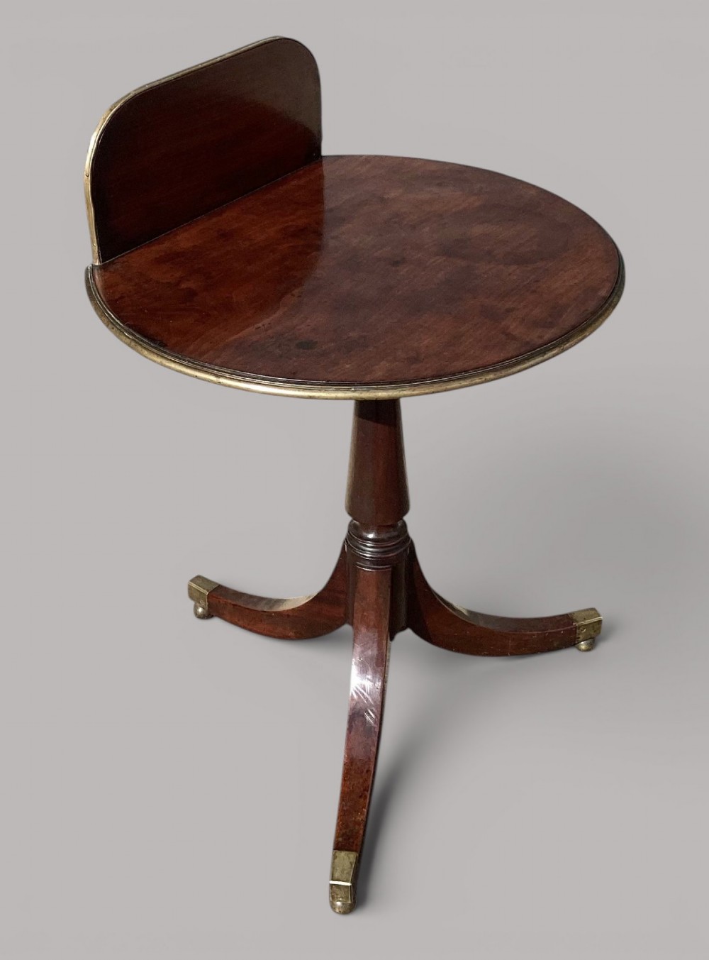 A George III mahogany tripod occasional table featuring a raised back panel, brass moulding on the circular top, a turned central column, and three curved legs ending in brass caps. View III