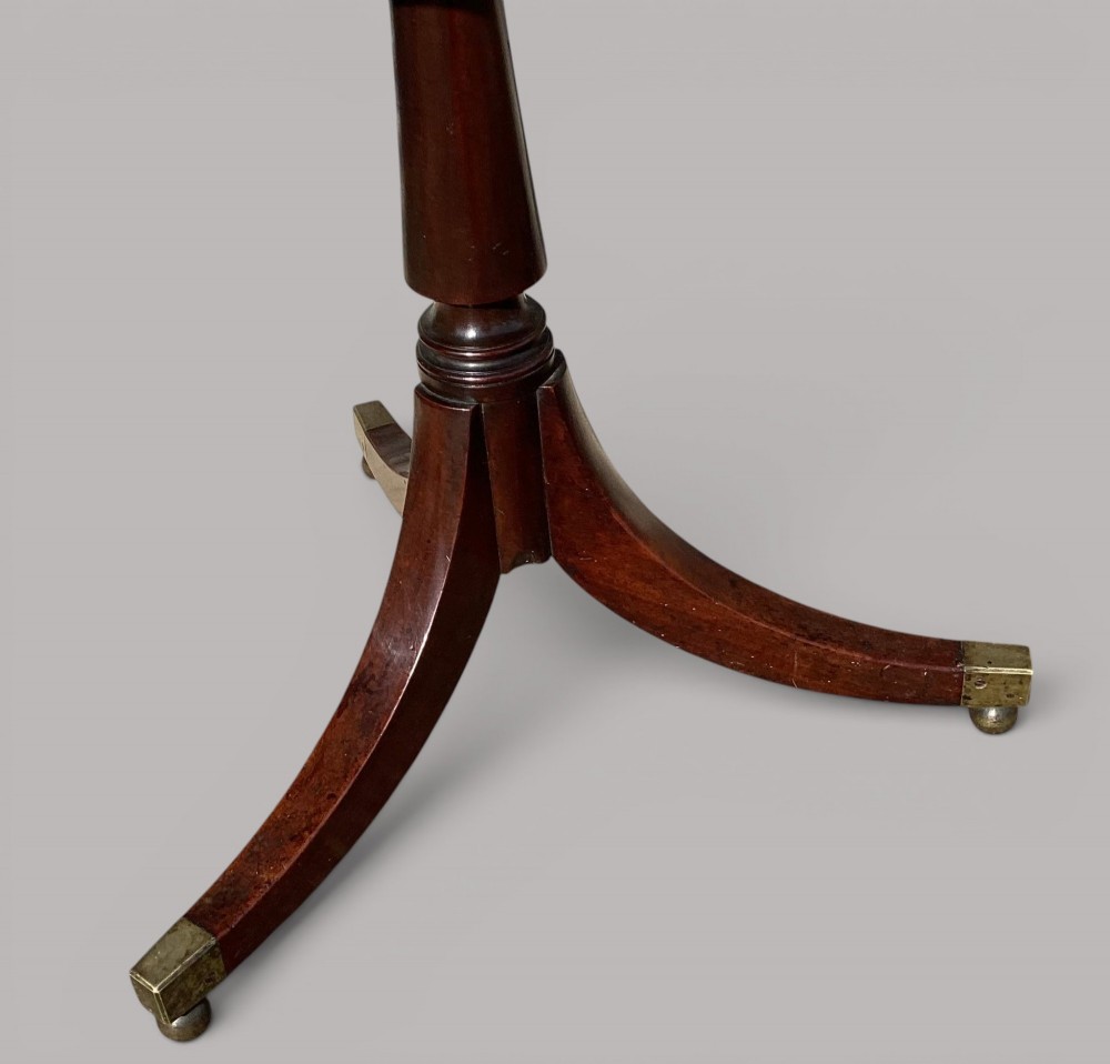 A George III mahogany tripod occasional table featuring a raised back panel, brass moulding on the circular top, a turned central column, and three curved legs ending in brass caps. View V