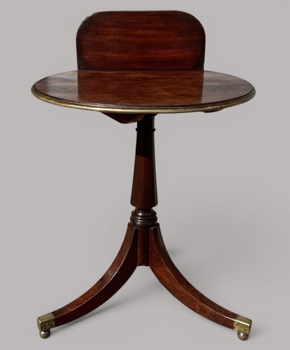 A George III mahogany tripod occasional table featuring a raised back panel, brass moulding on the circular top, a turned central column, and three curved legs ending in brass caps. View VI