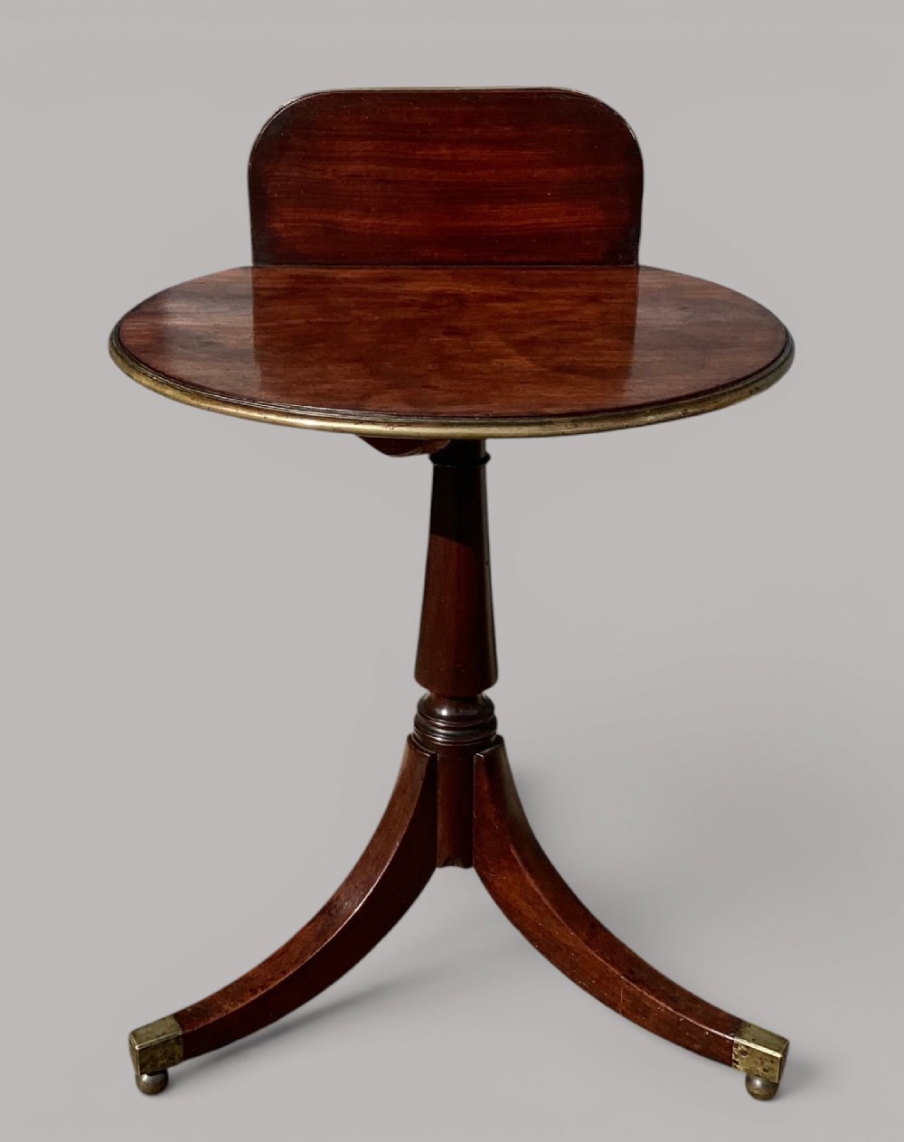 A George III mahogany tripod occasional table featuring a raised back panel, brass moulding on the circular top, a turned central column, and three curved legs ending in brass caps. View VII