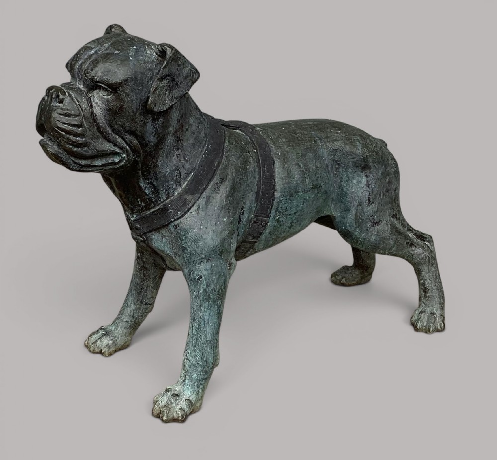 A life-size bronze sculpture of a bulldog standing while wearing a detailed harness, finished in a dark green verdigris patina.